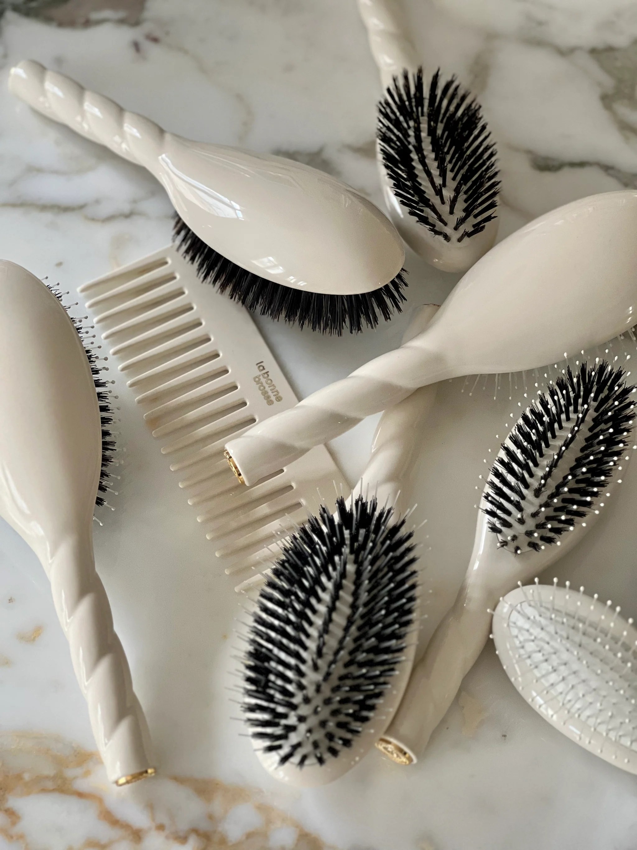 multiple hairbrushes and combs in an ivory color resting on a marble counter