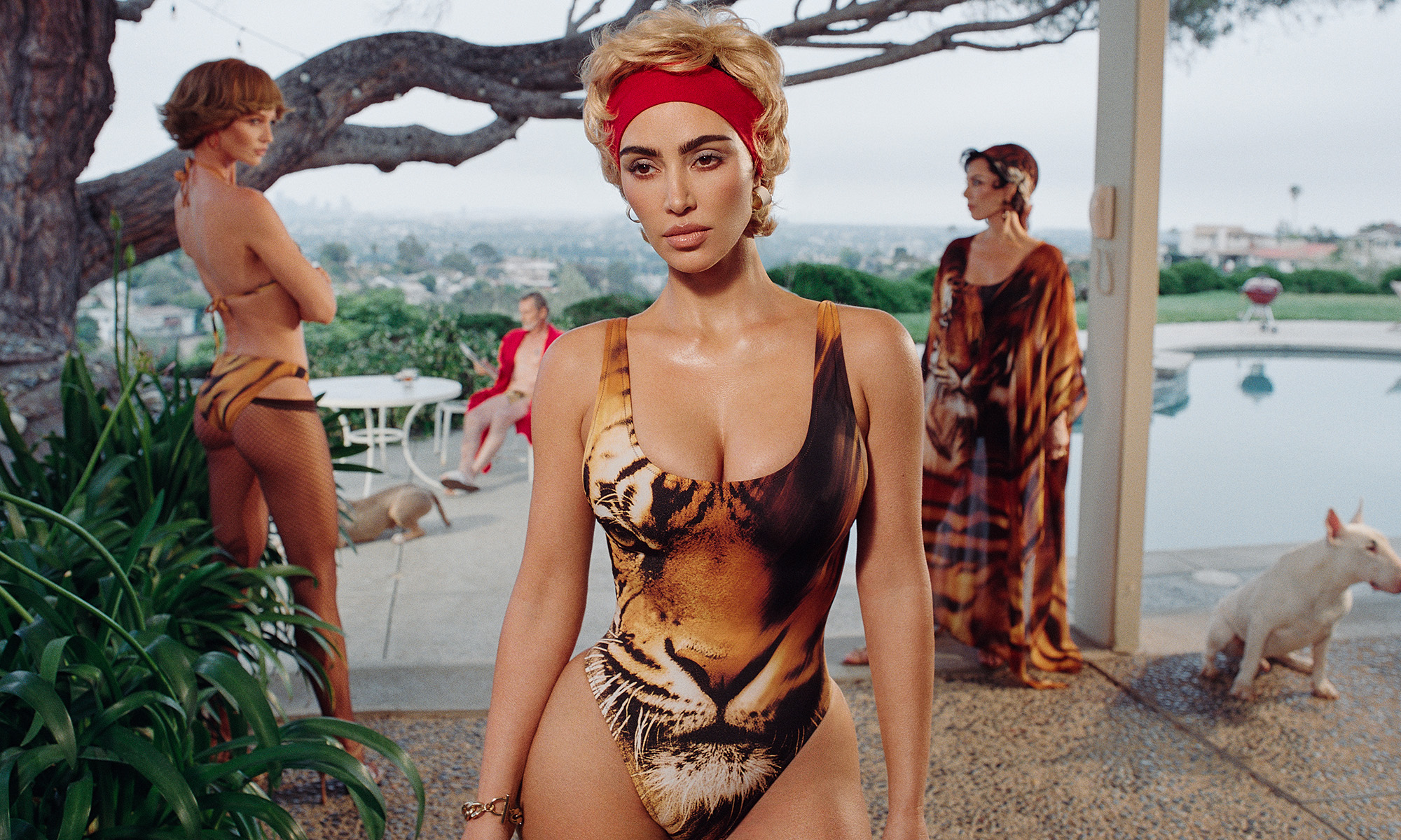 kim kardashian standing in a tiger-printed swimsuit with a red hairband, in front of two women and a dog outside